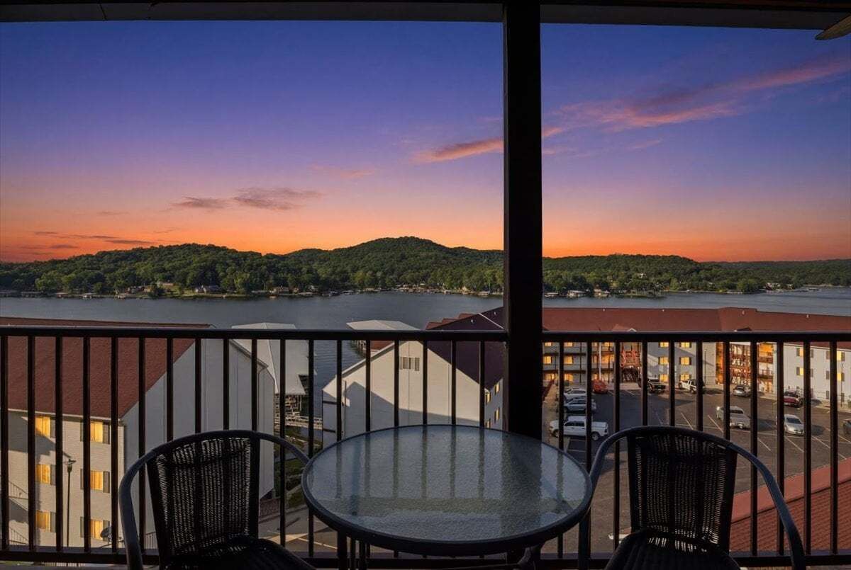 Un balcón con mesa y sillas de vidrio, con vistas a una masa de agua y un paisaje montañoso durante el atardecer.