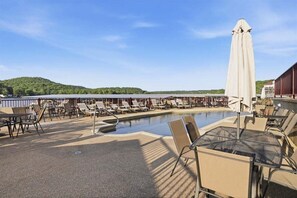 Zona de piscina con sillones reclinables, un espacio de descanso cubierto y vistas a un lago y colinas.