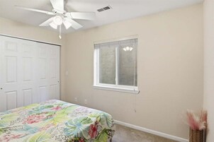 Un dormitorio con ventilador de techo, una ventana con persianas, una cama con colcha estampada y una pequeña cesta de mimbre.
