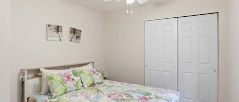 Un dormitorio con cama, ventilador de techo, una puerta y dos cuadros en la pared.