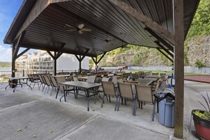 Un patio exterior con mesas y sillas, una zona cubierta con ventilador de techo y vistas a edificios al fondo.