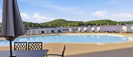 A swimming pool with lounge chairs and umbrellas, surrounded by a fence and hills in the background.