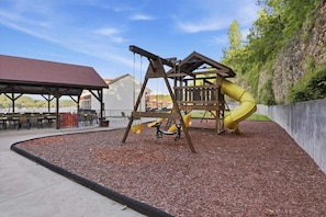 Un terrain de jeux équipé de balançoires et d’un toboggan, entouré d’un espace de repos abrité et d’un bâtiment doté d’un toit rouge.