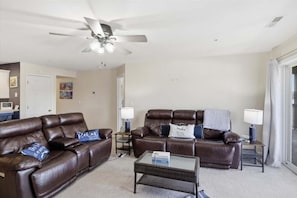 A living room with two brown leather sofas, a glass coffee table, a ceiling fan, and a door leading to another room. - Lakefront on Big Niangua, 2 Heated Pools, Boat Slip, Fire Pit, & Big Views (Camdenton)