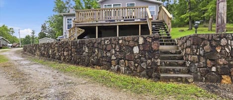 Una casa con un muro de piedra, una terraza de madera y una escalera que lleva a un segundo piso.