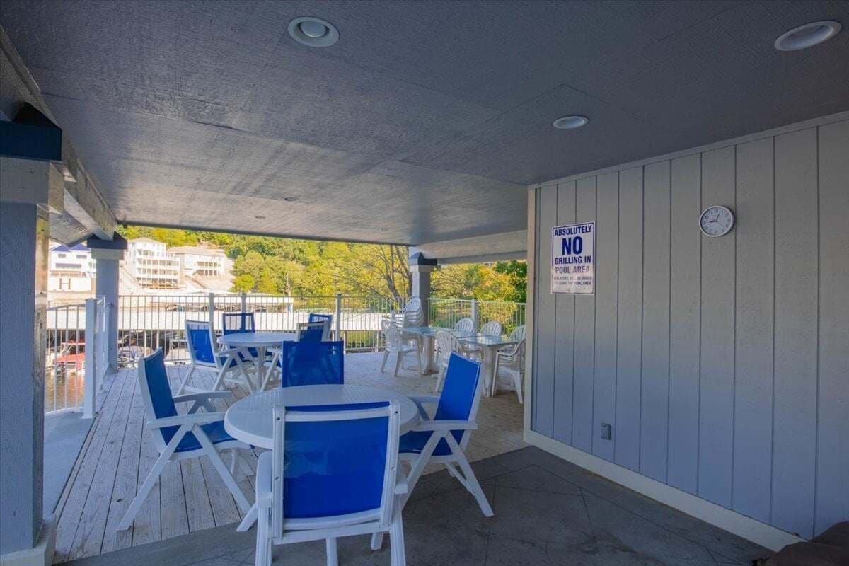 Un'area patio esterna con tavoli bianchi e sedie blu, una sveglia appesa al muro e un cartello che vieta l'uso del barbecue.