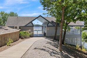 A building with a sloped roof, a gravel pathway leading to a gated entrance, and a view of a body of water in the background. - Top Floor Lake Views, Loft Space & Private Deck Retreat at the 12mm (Lake Ozark)