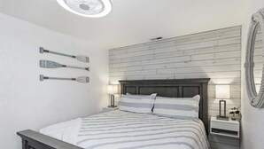 Une chambre à coucher comprenant un lit, deux tables de chevet, un miroir fixé au mur et un ventilateur de plafond.