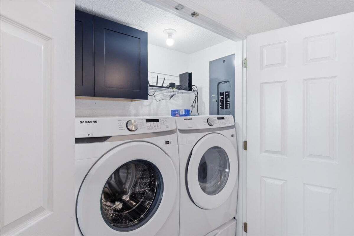 Un área de lavandería con dos lavadoras Samsung blancas, un mueble oscuro y una puerta blanca.