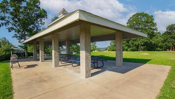 A covered picnic area with concrete pillars, picnic tables, and a barbecue grill.