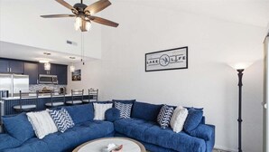 Un salon moderne avec un canapé d’angle, une table basse ronde, un ventilateur de plafond et une cuisine en arrière-plan.