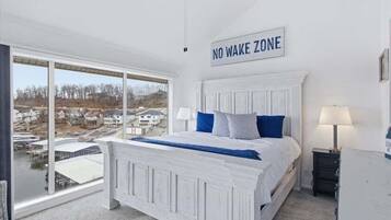 A bedroom with a large window, a bed with blue bedding, a 'NO WAKE ZONE' sign, and a view of houses and water.