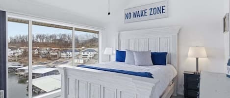 Un dormitorio con un ventanal grande, una cama con ropa de cama azul, un cartel que dice 'NO WAKE ZONE' y vistas a casas y agua.