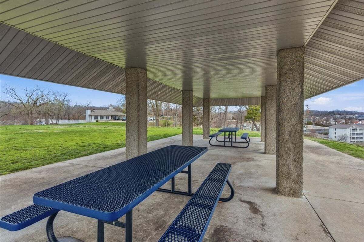An outdoor covered picnic area with benches and tables, surrounded by a grassy field and a building in the background.