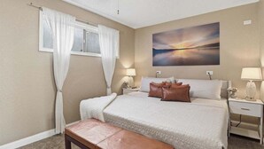 A bedroom with a bed, bedside tables, a bench, a window with curtains, a ceiling fan, and a painting on the wall.