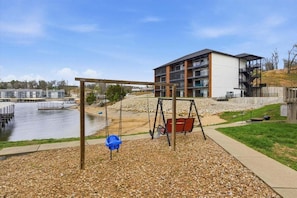 A playground with swings and a small blue bucket, a building with multiple floors and balconies, and a body of water in the background. - Bright Lakefront Condo, Amazing Views, Pools, Hot Tub (Osage Beach)
