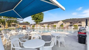 Zona de piscina al aire libre con sillas y mesas de plástico blanco, una sombrilla azul y un cielo despejado.