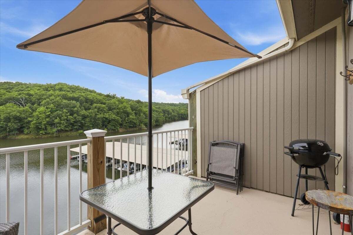 A balcony with a glass table, a barbecue grill, and a view of a lake.
