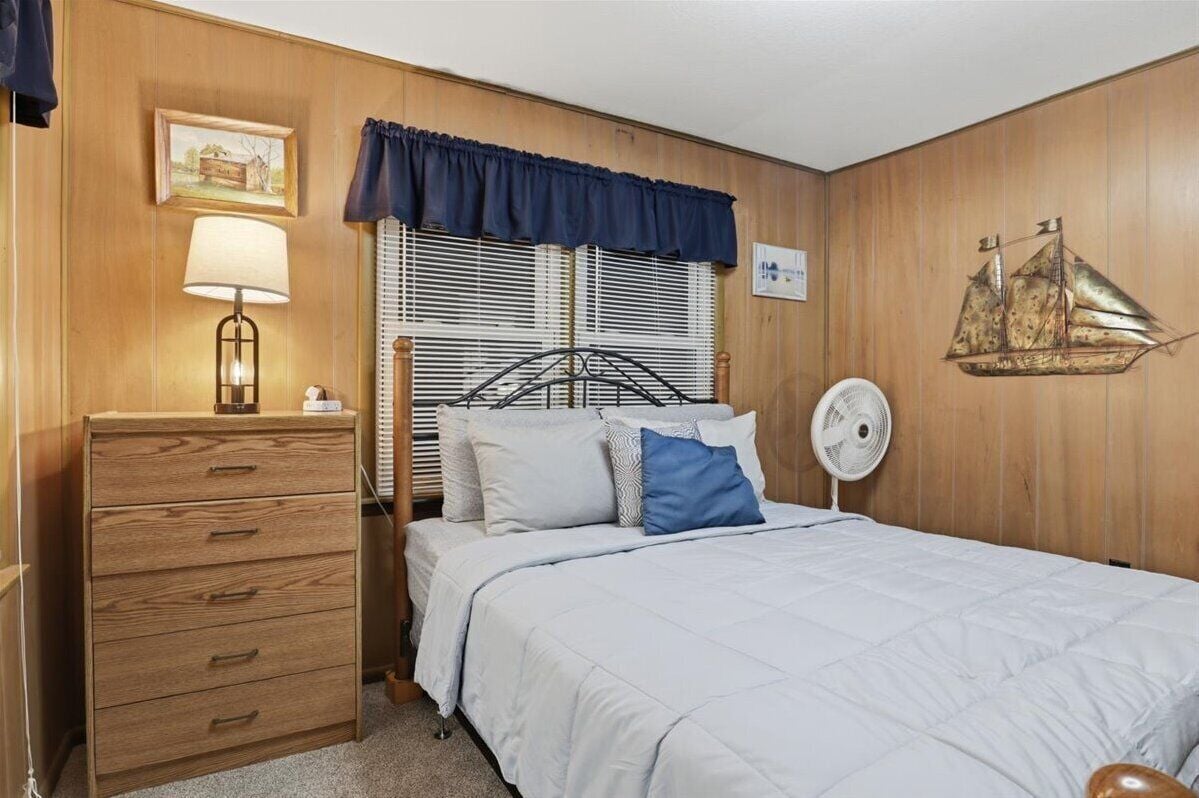 Un dormitorio con un aparador de madera, una cama con ropa de cama blanca, una almohada azul, un ventilador de pared y una decoración náutica.