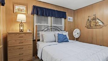 Un dormitorio con un aparador de madera, una cama con ropa de cama blanca, una almohada azul, un ventilador de pared y una decoración náutica.