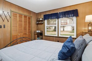 Quarto com cama, janela e vista, porta de madeira e prateleira com objetos.