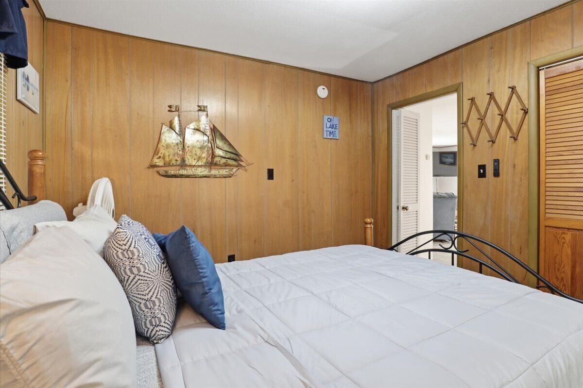 A bedroom with wooden paneling, a bed with white bedding, patterned pillows, a wall-mounted ship decoration, a door with a sign, and a wall-mounted light fixture.