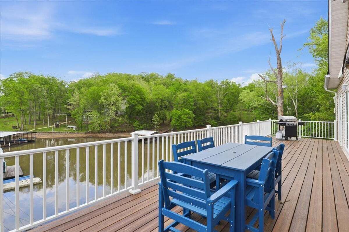 Une terrasse en bois avec une table et des chaises, un barbecue et une vue sur un lac bordé d’arbres.