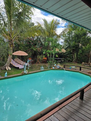 A swimming pool with a wooden deck, surrounded by palm trees and a thatched roof structure.