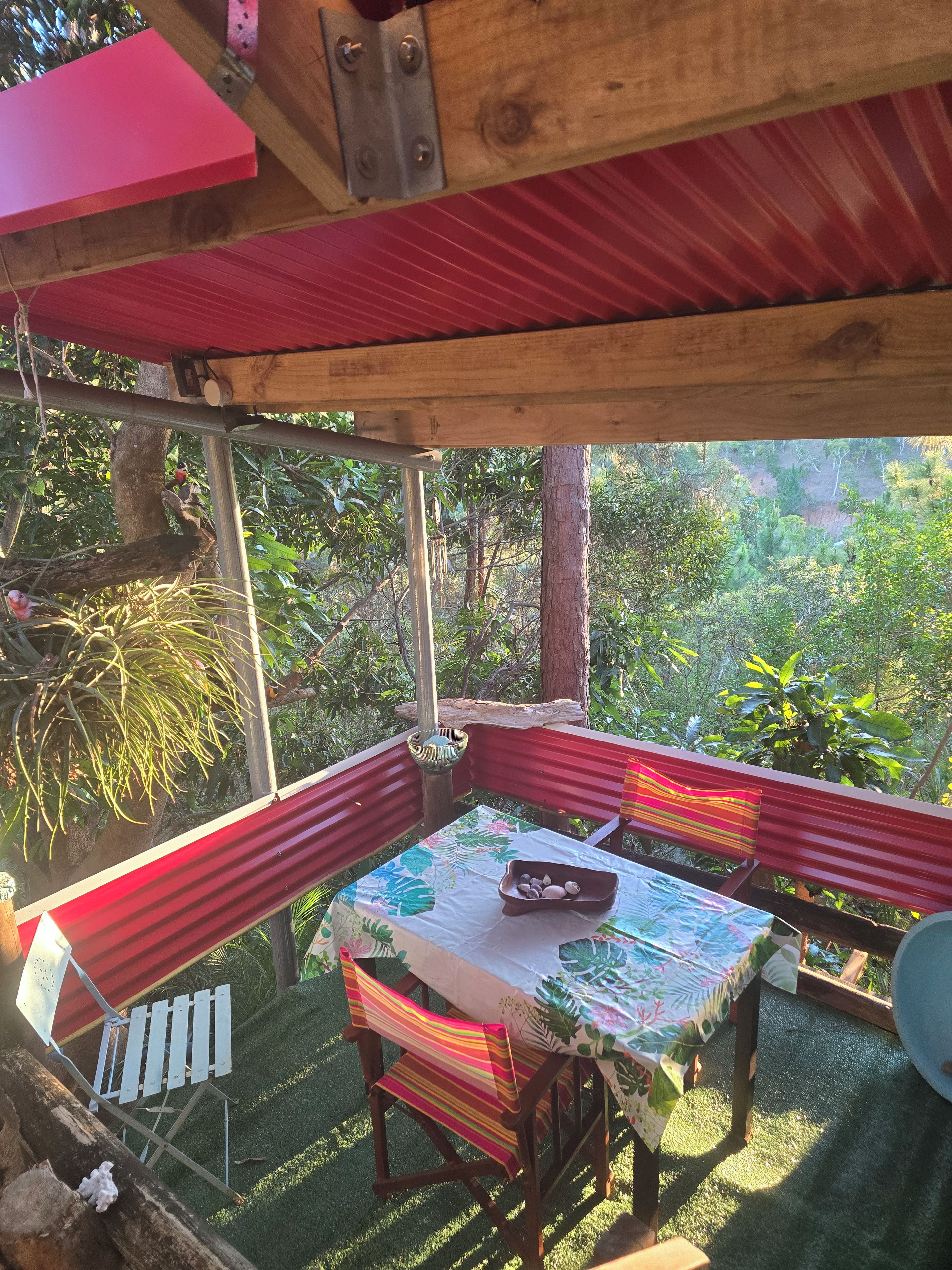 A covered outdoor seating area with a table and chairs, surrounded by greenery.