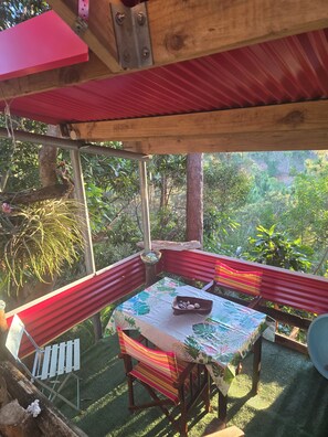 A covered outdoor seating area with a table and chairs, surrounded by greenery. - Gîte du tour du monde (Païta)
