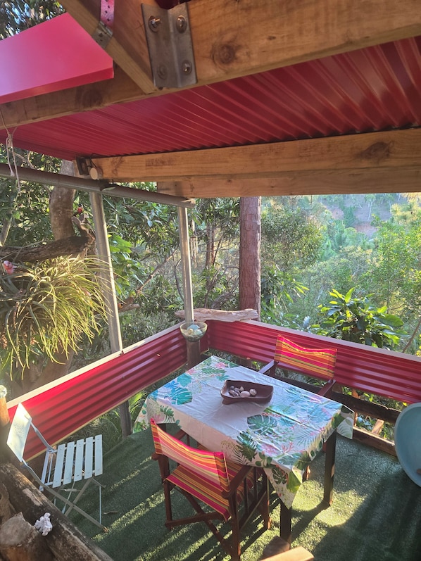 A covered outdoor seating area with a table and chairs, surrounded by greenery.