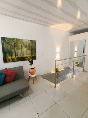 A modern living room with a grey sofa, a large forest-themed painting, a potted plant, and a staircase with a glass railing.