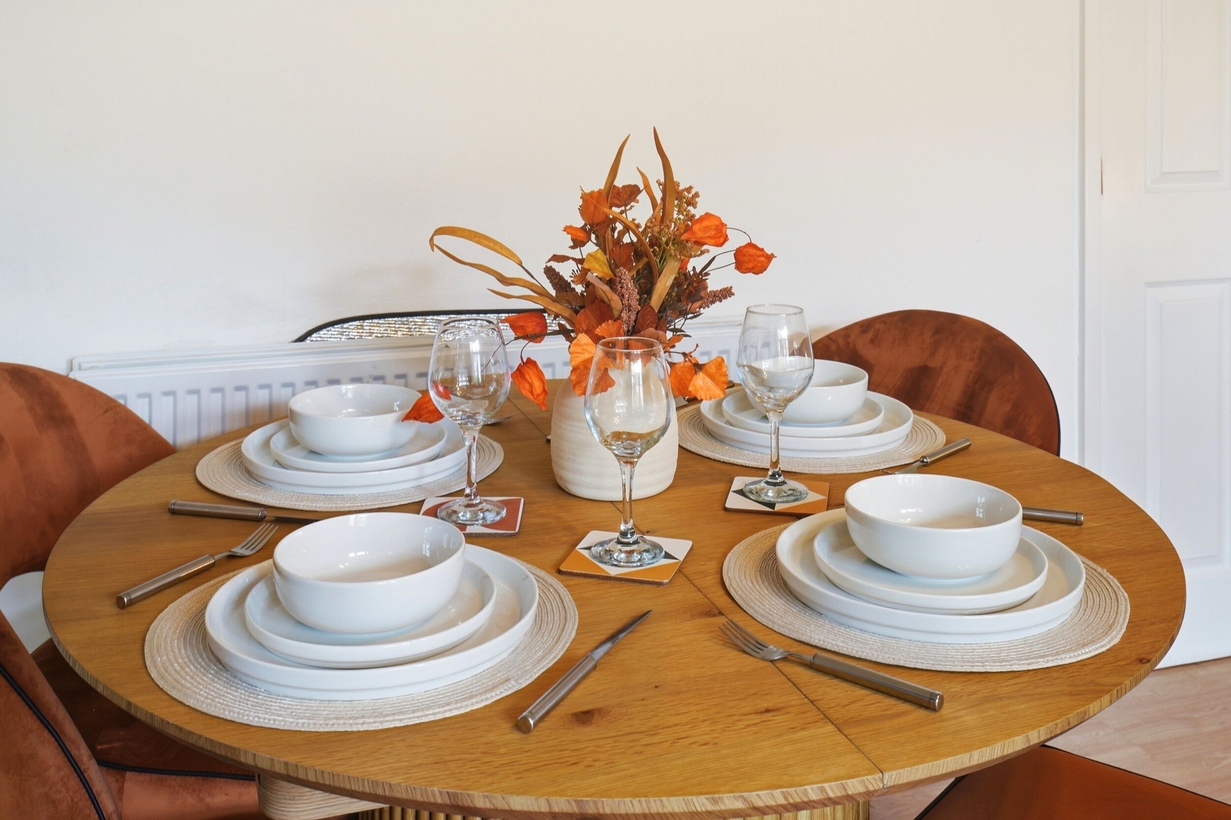 A round dining table set with white plates, bowls, and cutlery, accompanied by a vase of autumn flowers.