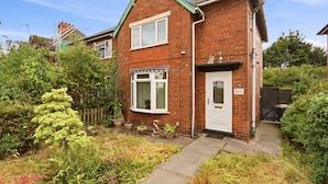 A two-story brick house with a white door and window shutters, surrounded by a garden with various plants and a wooden fence. - Crew Comfort Walsall | Sleeps 6 | Fast Wi-Fi (Walsall)