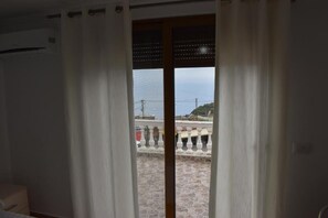 A room with a view through a window with curtains, a stone floor, and a white railing outside. - Kompleks Angjelo (Lukovë)