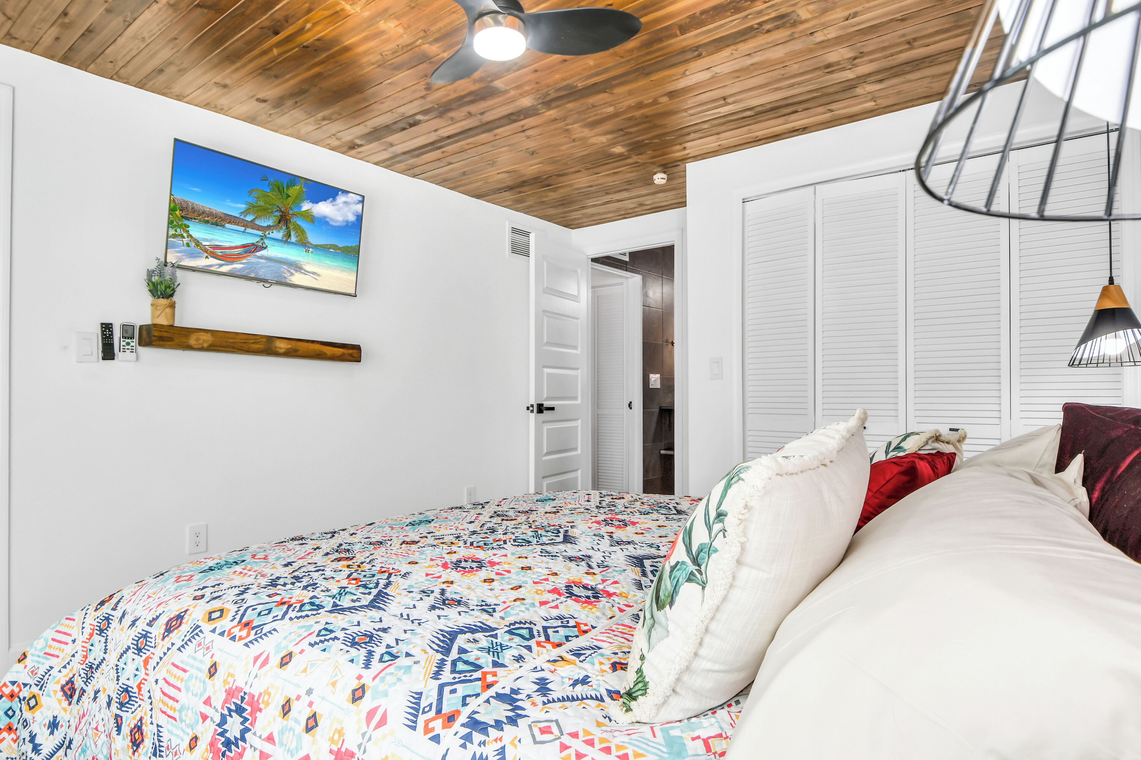 Une chambre à coucher avec un lit, un tableau représentant une scène de plage, un ventilateur de plafond et une porte donnant sur une autre pièce.