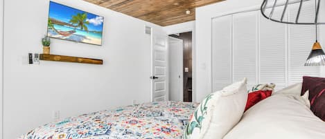 Un dormitorio con una cama, un cuadro de una escena de playa, un ventilador de techo y una puerta que da a otra habitación.