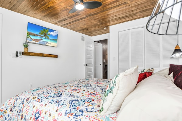 A bedroom with a bed, a painting of a beach scene, a ceiling fan, and a door leading to another room.