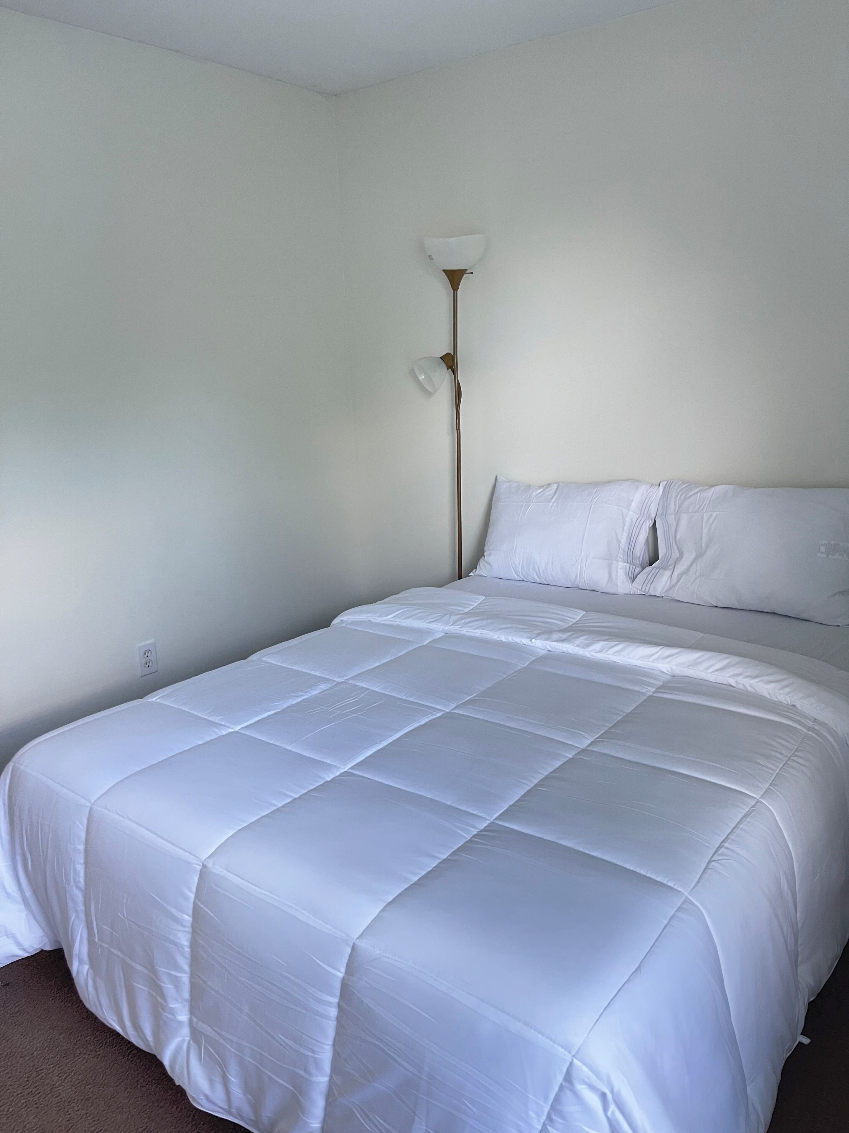 A bedroom with a bed, white bedding, a floor lamp, and a plain wall.