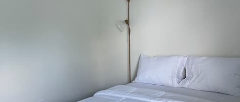 Un dormitorio con cama, ropa de cama blanca, una lámpara de pie y una pared lisa.