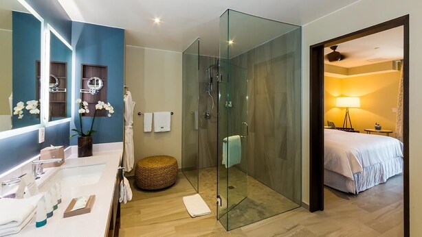 Un baño moderno con ducha de vidrio, tocador con espejo y una habitación separada visible a través de una puerta abierta.