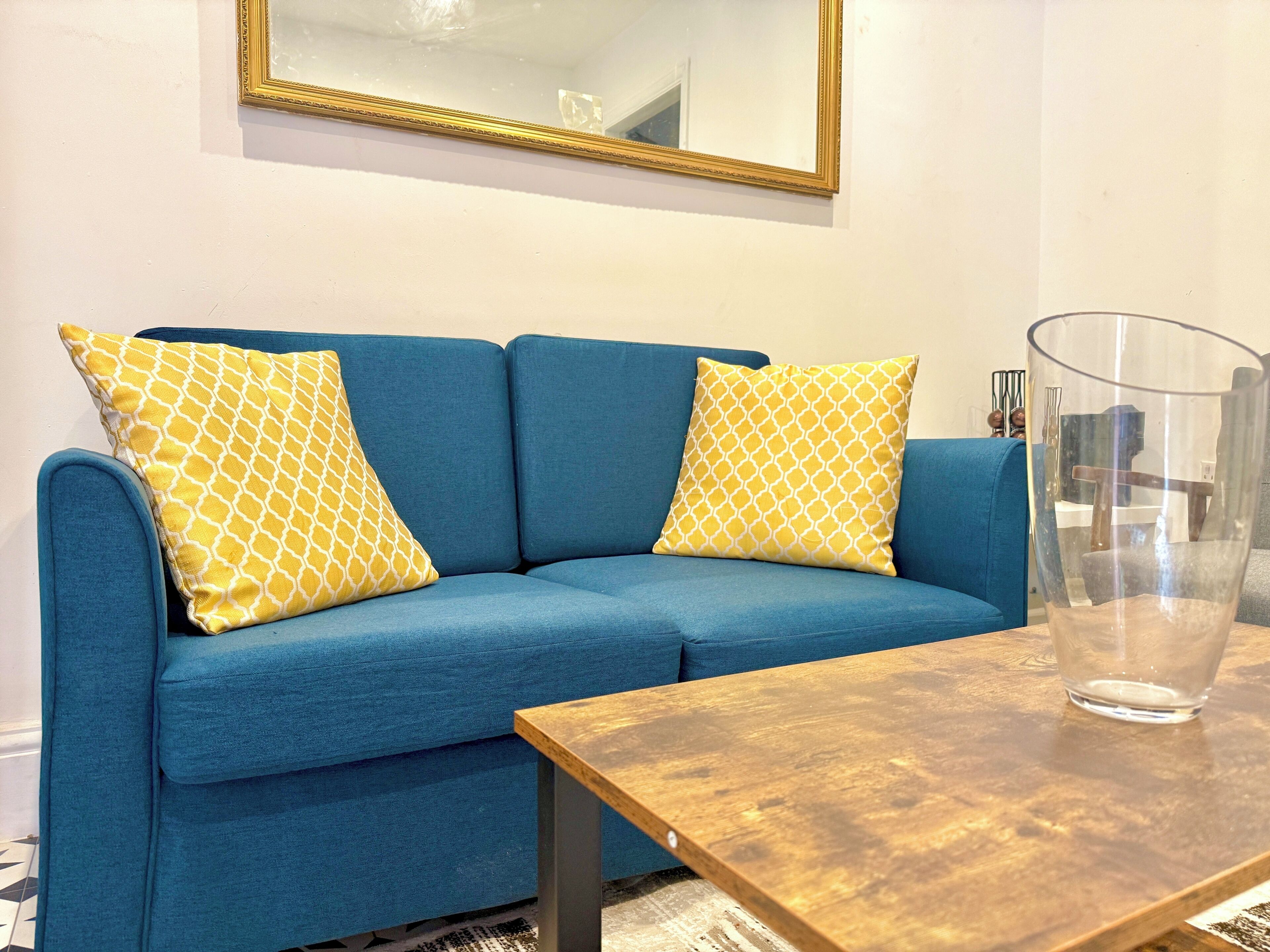 A blue sofa with patterned cushions, a wooden table, and a clear glass vase.