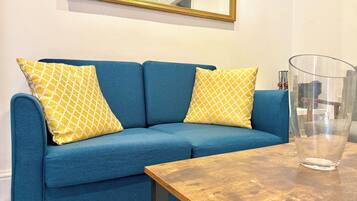 A blue sofa with patterned cushions, a wooden table, and a clear glass vase.