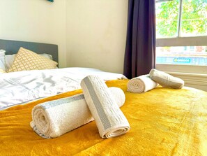 Una cama con una manta amarilla, dos toallas enrolladas y una almohada estampada.