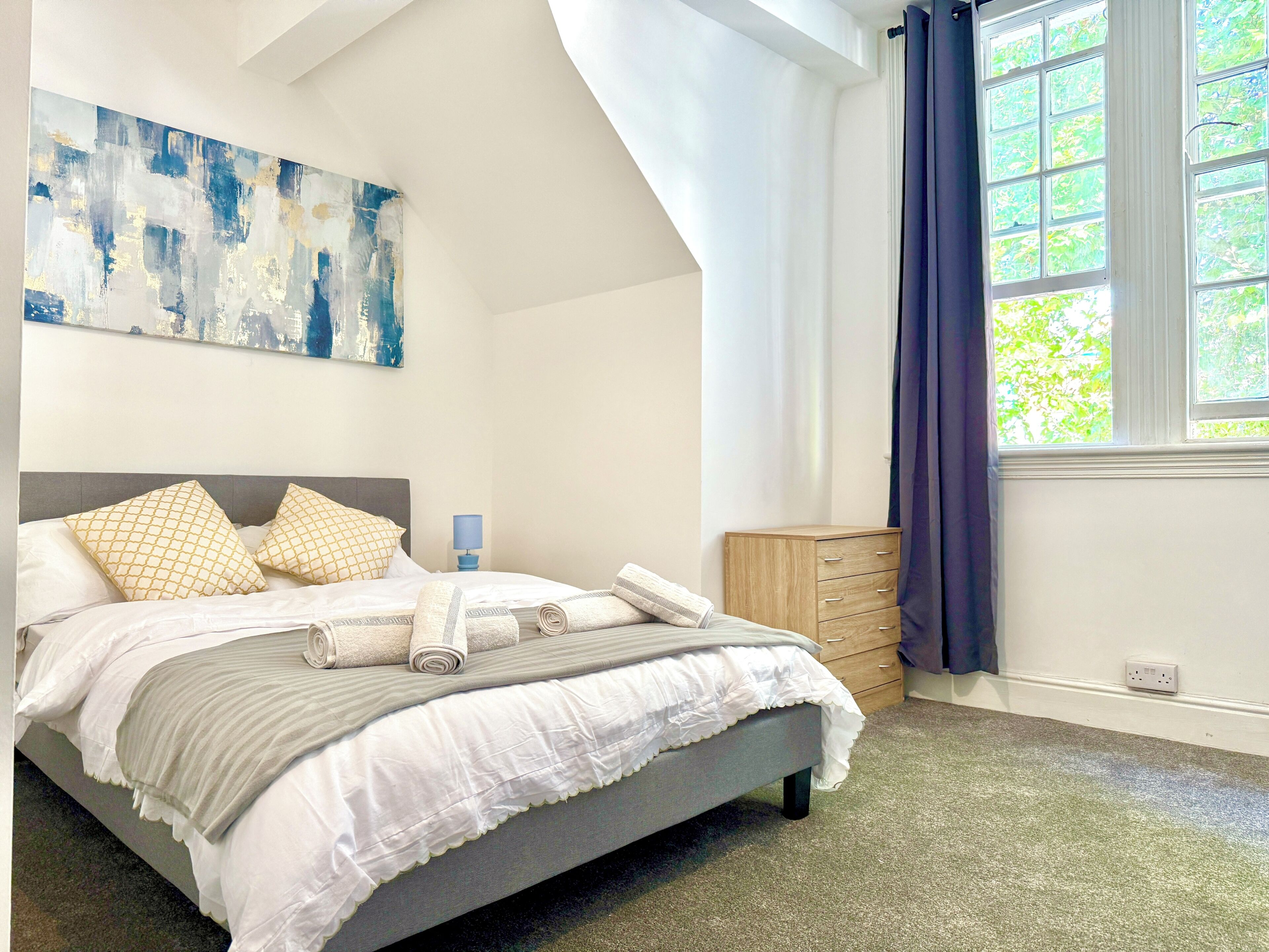 A bedroom with a bed, a nightstand, a dresser, a window with curtains, and a painting on the wall.