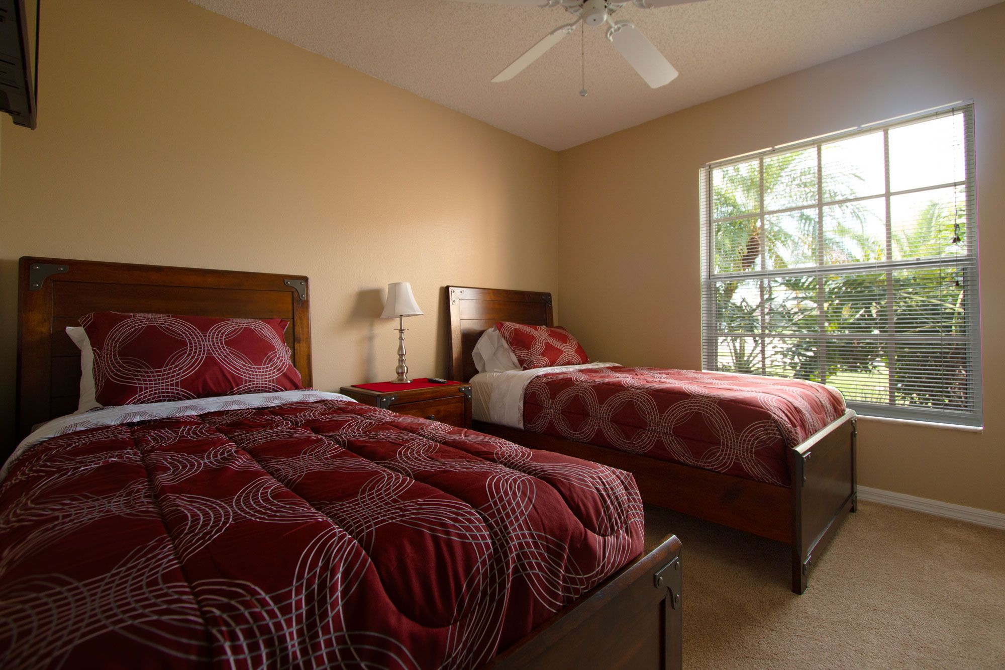 Quarto de hotel com duas camas, um ventilador de teto, uma mesa de cabeceira com abajur e uma janela com persianas.