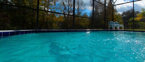 A swimming pool with clear blue water, surrounded by trees and a covered area with chairs.