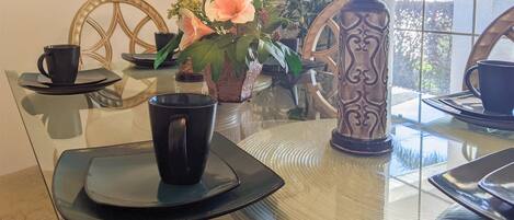 A glass-top dining table with black coffee cups and saucers, a decorative vase with flowers, and a wooden chair with a woven backrest.