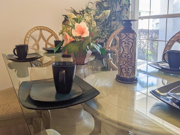 Ett matbord med glasskiva, svarta kaffekoppar och tillhörande fat, en dekorativ vas med blommor och en trästol med ett flätat ryggstöd.