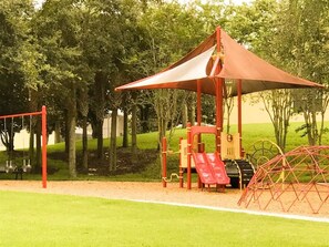 Ein Spielplatz mit rotem Sonnensegel, Schaukeln und Rutschen.